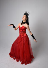Full length  portrait of beautiful young asian woman wearing red corset and ornate gothic queen crown. Graceful standing posing  isolated on studio background.