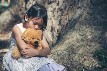 Sad girl hugging teddy bear sitting under tree sadness alone in green park. Lonely girl feeling sad...