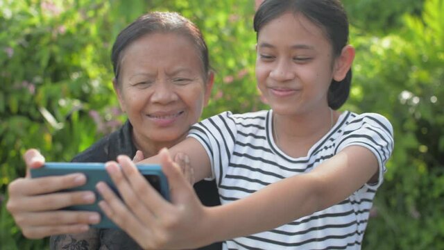 Cheerful adorable teen granddaughter and her senior grandmother taking selfie photo with mobile smartphone and smiling while looking at camera in the garden at home. Bonding relationship in family.