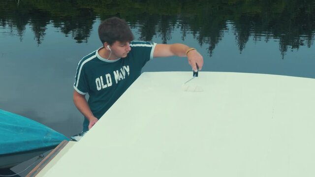 Young man finishes thinned coat of white primer on wooden boat roof