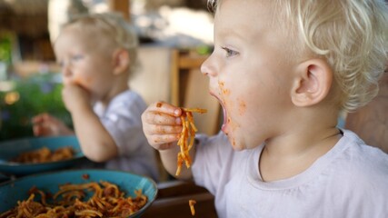 Side view of two white babies learning to feed themselves with plant based pasta in an outdoor restaurant. Messy skill developing