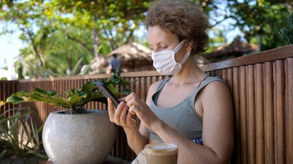 Young woman in a face mask relaxing in an tropical garden drinking latte coffee and browsing on her phone