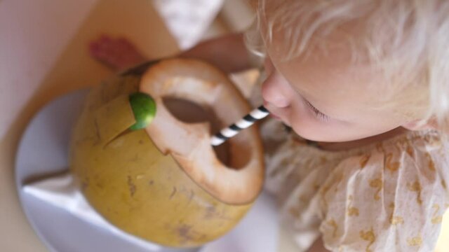 Top view of toddler baby girl at tropical vacation drinking green young coconut with carved heart on it from paper straw