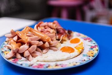 Breakfast, sausages, fried eggs and bacon wrapped cheese in the same plate, laying on a blue table.