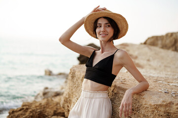 Portrait of woman in black bralette and white trousers leaning on rock at beach