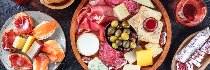 Charcuterie and cheese platter panorama with rose wine and salmon sandwiches, top shot on a black...
