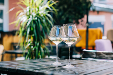 A wooden table in a restaurant with white wine. Wine glasses served for a party in a bar or a restaurant on terrace at a sunny day. Blurred background with plants. Still life scene