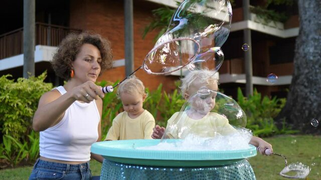 Creating childhood memories. Caucasian mother and her twin toddler daughters play with foam bubbles. Palm trees in the background 