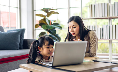 Mother and asian kid little girl learning and looking at laptop computer making homework studying knowledge with online education e-learning system.children video conference with teacher tutor at home