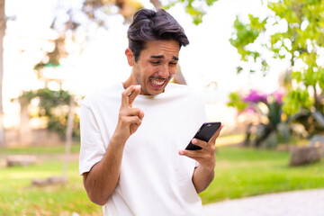 Fototapeta premium Young caucasian man in a park using mobile phone with fingers crossing