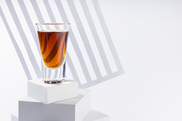 Bright amber brandy cocktail in shot glass in sunlight on white square podium as stairs with diagonal abstract stripes in soft light space in luxury minimal geometric style.