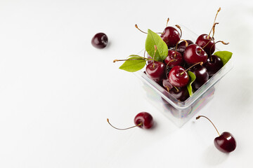 Ripe red fresh wet cherry in plastic box with green leaves, water drops and tail in sunlight on white wood board, top view, copy space.