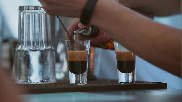 Bartender prepares cocktail B52 on table. Man holds spoon and carefully pours third transparent layer of liquor. Preparing three-layer cocktail for nightclub party. Light is changing.