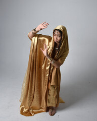 Full length portrait of pretty young asian woman wearing golden Arabian robes like a genie, standing pose holding flowing fabric, isolated on studio background.