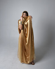 Naklejka premium Full length portrait of pretty young asian woman wearing golden Arabian robes like a genie, standing pose holding flowing fabric, isolated on studio background.