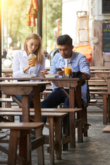Young woman drinking refreshing cocktail at outdoor cafe and looking at boyfriend texting friends or using app on smartphone