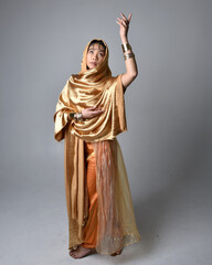 Full length portrait of pretty young asian woman wearing golden Arabian robes like a genie, standing pose holding flowing fabric, isolated on studio background.
