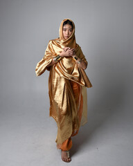 Full length portrait of pretty young asian woman wearing golden Arabian robes like a genie, standing pose holding flowing fabric, isolated on studio background.