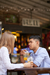 Pretty smiling young woman enjoying refreshing drink and conversation with boyfriend when sitting at tablet in outdoor cafe