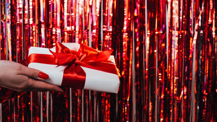 Christmas greeting. Holiday gift. Party celebration. Female hands showing gift box atlas ruby ribbon on shimmering sparkle red cascade curtain