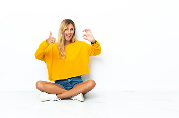 Young blonde Uruguayan woman sitting on the floor isolated on white background showing ok sign and...