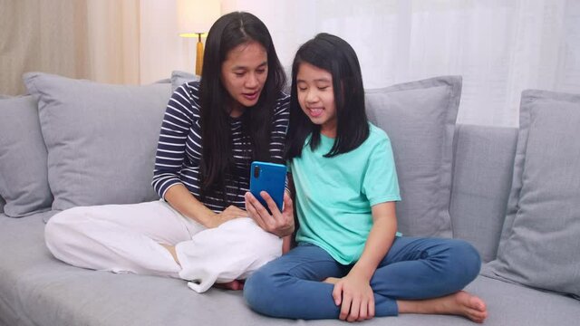 Happy Asian family mother and daughter sitting on sofa talking video conference by smartphone together in living room at home. mother and cute little girl video call by smartphone.Technology lifestyle
