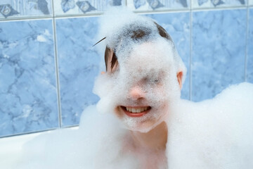 Portrait of happy child boy is playing with white foam sitting in bath at home, water games. Sensory games for children's development. He makes hat from foam on head. Looking at camera and smiling.