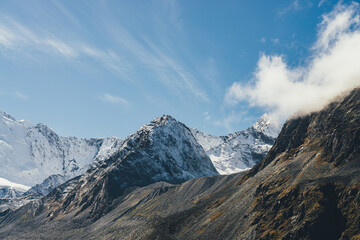 Atmospheric alpine landscape with high snowy mountain with peaked top under cirrus clouds in sky. Big snow covered mountain in sunshine. Low cloud on black rocks and white-snow pointy peak in sunlight