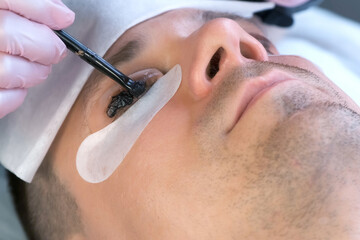 Cosmetologist puts black paint on the man's lashes using brush. Laminating eyelashes procedure in...