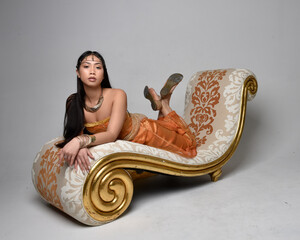 Full length portrait of pretty young asian woman wearing golden Arabian robes like a genie, seated...