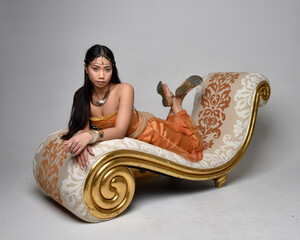 Full length portrait of pretty young asian woman wearing golden Arabian robes like a genie, seated...