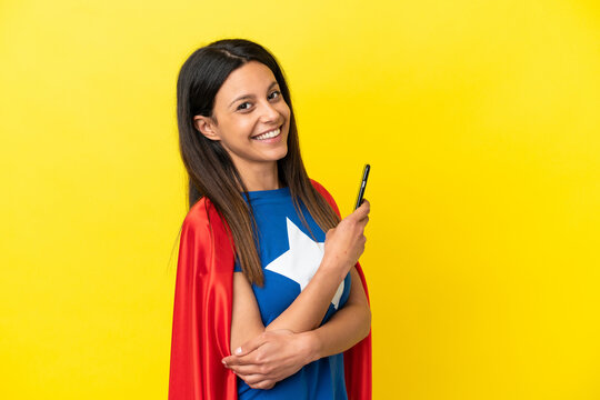 Super Hero woman isolated on yellow background holding a mobile phone and with arms crossed