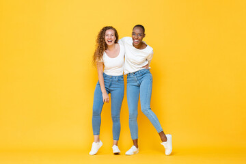 Happy two interracial woman friends smiling and holding each other in isolated studio yellow color...