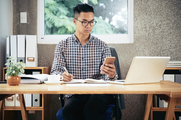 Fototapeta premium Serious young entrepreneur in plaid shirt working at office desk, he is reading text messages and taking notes in planner