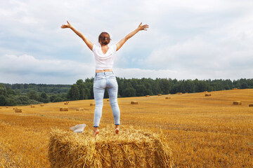 girl with straw hat stands on a haystack on a bale in the agricultural field after harvesting