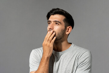 Shocked young Caucasian man looking up and thinking with hand cover mouth on light gray studio...