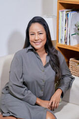 Positive mature woman in grey shirt resting in armchair and smiling at camera