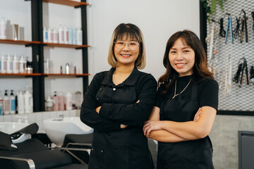 Portrait of team of happy successful young Asian hairstylist standing wearing apron and spectacles with folded hands smiling while looking at camera
