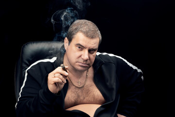 an adult white man in a tracksuit is sitting in a chair and smoking a cigar, looking at the camera....