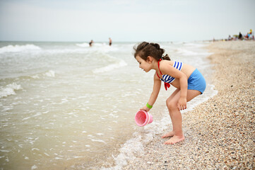 Pretty baby girl in a swimsuit collecting sea water in a pink toy bucket. Cute child playing on the beach. Healthy rest, recovery and recreation of children during the summer holidays.