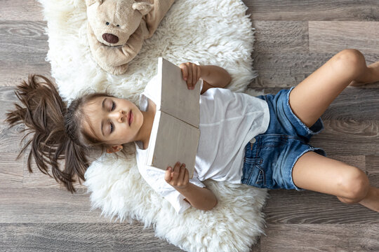 Little girl reads a book lying on the floor at home, top view.
