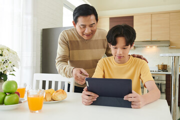 Happy father patting his son on shoulder and looking at screen of tablet computer with educational...
