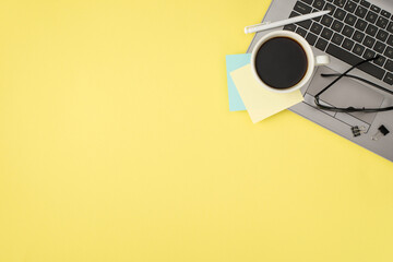 Top view photo of cup of coffee yellow and blue sticker note paper black glasses binder clips and white pen on laptop on isolated pastel yellow background with blank space