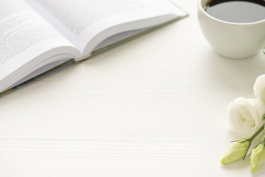 Photo of white cup of coffee flowers and open book on white table with copyspace