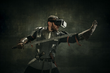Portrait of one brutal bearded man, medeival warrior or knight in VR glasses with sword over dark background.