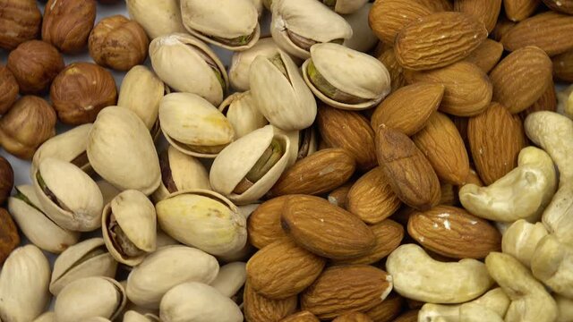 The roasted pistachio seeds with shell, blanched almonds, hazelnuts and cashew nuts lie in rows on a white table. Assorted different nuts, background.