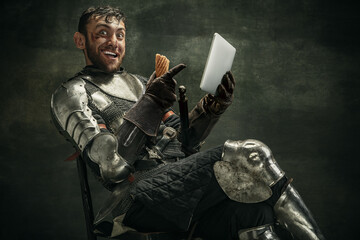One brutal bearded man, medeival warrior or knight with digital tablet sitting on chair over dark...