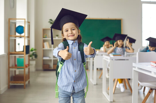 Schoolboy is wearing a graduation cap, showing thumb up. Schoolboy stands on blurred background of class, graduation, school board, back to school. Smart elementary school kid