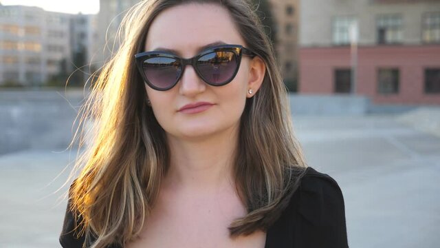 Portrait of young businesswoman in sunglasses walking in city street. Face of attractive business woman looking at camera. Sunset light illuminates hair of girl. Close up Slow motion