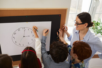 Primary students learning to tell the time. Young female teacher and group of happy elementary school children standing in front of modern classroom board and working with clock poster all together